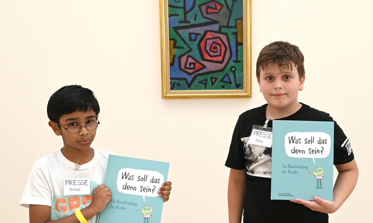 Zwei Jungs im Museum, halten beide einen Kunstkatalog in den Händen „Was soll das denn sein?“, im Hintergrund ein Gemälde