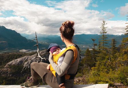 Frau mit Baby im Tragegurt, blickt auf Bergkulisse