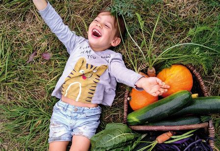 Ein Mädchen liegt lachend auf einer Wiese, neben ihr ein Korb mit Gemüse