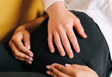 Close-up eines Schwangerschaftsbauches, auf dem drei Hände ruhen, schwarze Hose, weißes Shirt im Hintergrund gelbes Sofa
