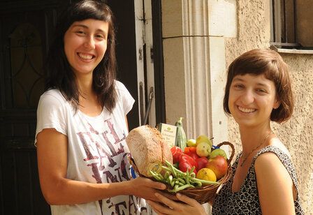 Zwei Frauen stehen mit einen Korb, in dem Obst, Gemüse, Brot und Tee ist, vor einer Haustür und lächeln in die Kamera