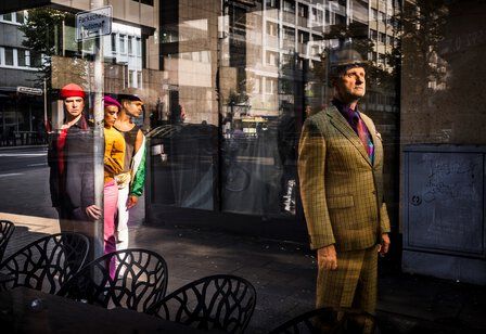 Ein Mann mit Melone und kariertem Anzug steht vor einem Schaufenster, hinter seinem Rücken blicken drei Kinder auf ihn.