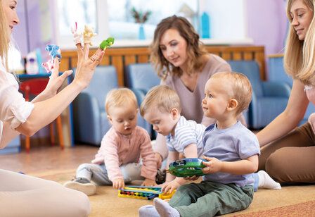 Drei Betreuerinnen und drei Kleinkinder sitzen auf dem Boden spielen zusammen, eine Betreuerin hat Fingerpuppen auf den Fingern