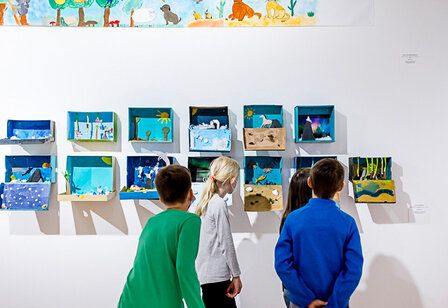 Kinder stehen vor selbstgemachten Kunstwerken, die an der wand hängen und betrachten die