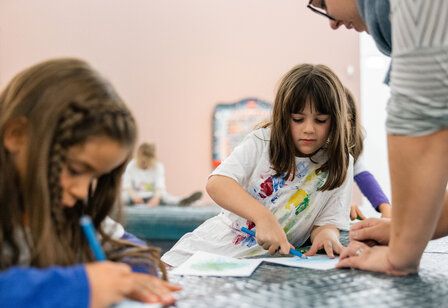 Kinder sitzen zusammen und zeichnen, eine Frau spricht mit einem Mädchen