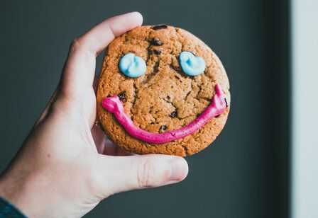 Hand hält einen Keks in die Kamera, der ein lustiges Gesicht aus Zuckerguss hat