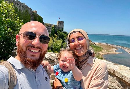 Vater und Mutter mit Baby vor einer bewachsenen Ruine am Meer