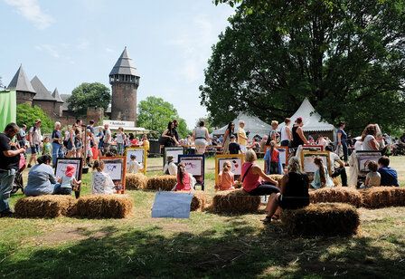 Leute sitzen auf Heuballen und malen Bilder, im Hintergund Marktstände und die Burg Linn