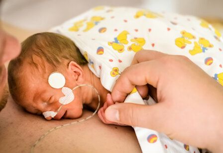 ein Frühchen mit einem Schlauchen in der Nase liegt auf der Brust des Vaters, er hält die kleine Hand fest