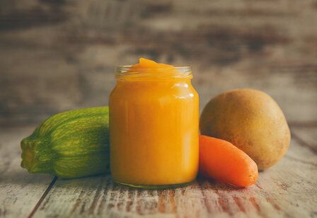 ein Gläschen mit Gemüsebrei steht auf einem Holztisch, daneben liegen eine Zucchini, eine Mähre und eine Kartoffel