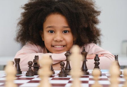 Mädchen hinter Schachbrett, den Kopf auf die verschränkten Arme gestützt, lächelt in die Kamera, im Vordergrund Schachfiguren, teilweise unscharf