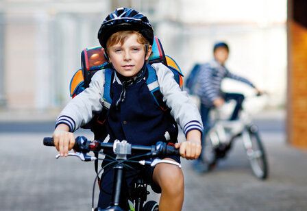 Junge auf Fahrrad, mit Helm und Tornister, anderer Junde auf Fahrrad im Hintergrund unscharf