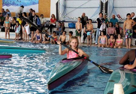 Schwimmhalle, viele Kinder sitzen oder stehen am Beckenrand, ein Mädchen paddelt in einem Boot