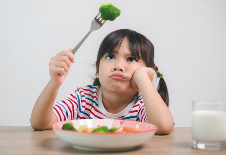 kleines Mädchen hat ein Brokkoliröschen auf der Gabel und guckt es trotzig an