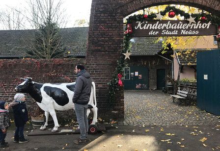 Vor dem Eingangstor des Kinderbauernhofs Neuss steht die Figur einer Kuh, ein Vater und zwei Kinder betrachten sie, der Eingang ist weihnachtlich geschmückt