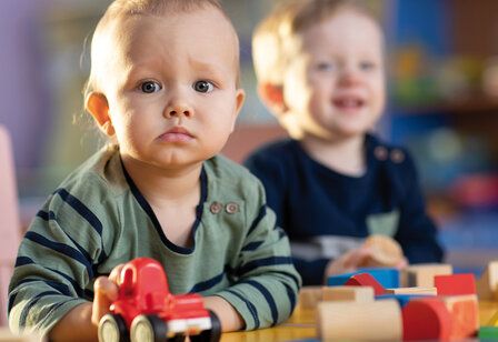 Kleinkind spielt mit Holzspielzeug und guckt skeptisch in die Kamera, im Hintergrund ein lachendes Kleinkind, unscharf