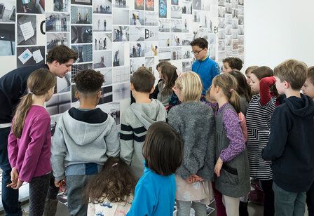 Museumspädagoge steht mit Kindern vor einem Kunstwerk und spricht mit ihnen darüber