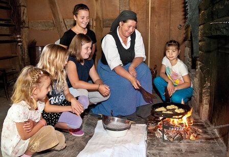 Frau in traditioneller Tracht brät etwas in einer Pfanne auf dem Boden eines Hauses im Freilichtmuseum Kommern, fünf Mädchen gucken zu