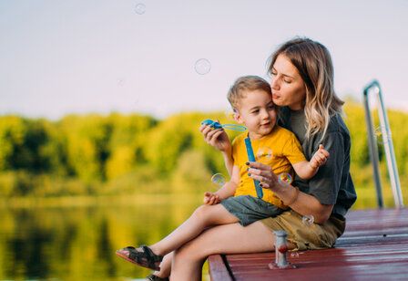 Eine Mutter sitzt mit ihrem Kind auf einem Steg an einem See und pustet Seifenblasen