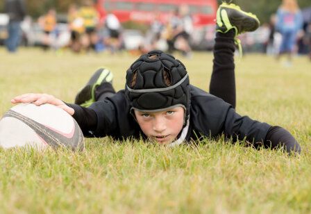 ein junger Rugbyspieler liegt auf dem Rasenplatz, guckt in die Kamera und hält mit einer Hand den Ball fest