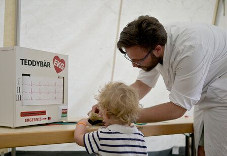 Arzt untersucht einen Teddybären, kleines Kind schaut zu