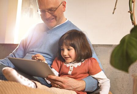 Opa sitzt mit Enkelin zusammen und beide schauen aufs Tablet.