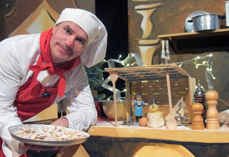 Mann mit Kochmütze und Schürze zeigt einen Teller mit Keksen, hinter ihm eine Küchen-Kulisse