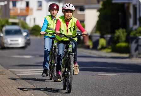 Zwei Kinder mit Helm und Warnweste fahren au einer Straße Fahrrad