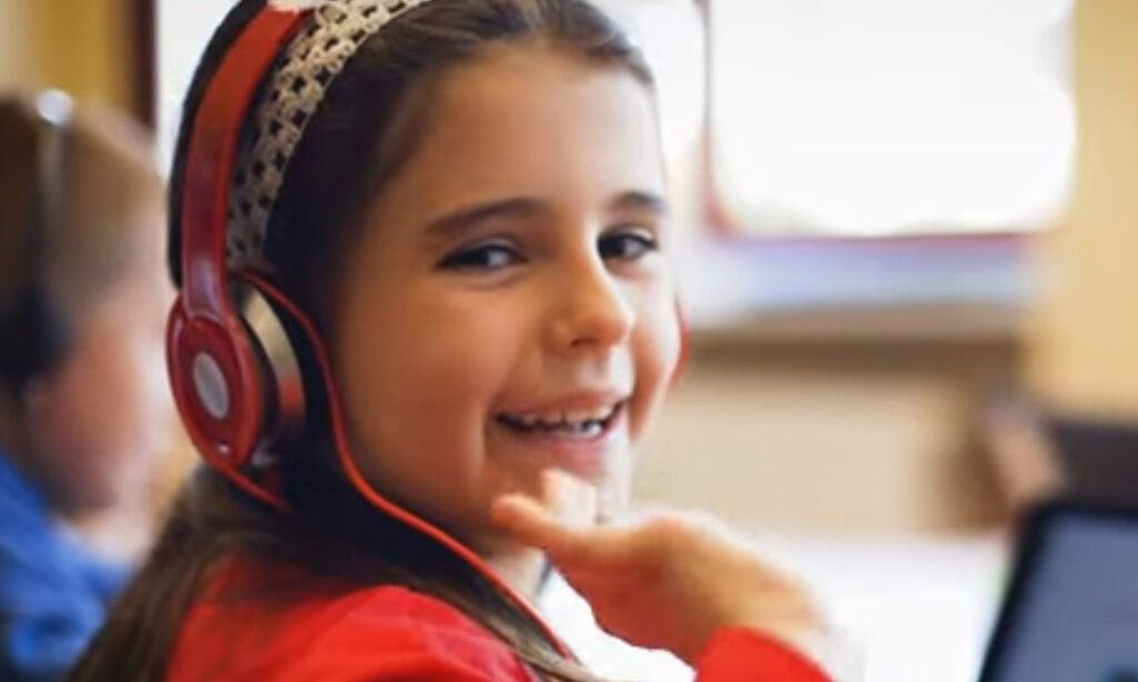 Mädchen im Klassenzimmer mit rotem Kopfhörer winkt lachend
