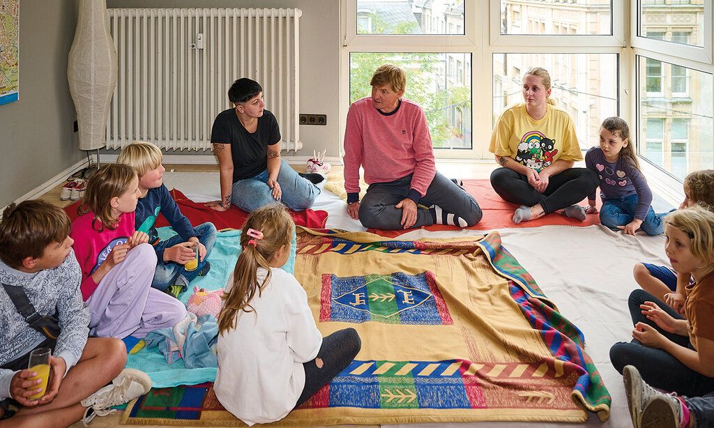 Mehrere Kinder und drei Erwachsene sitzen in einem Raum auf dem Boden und reden miteinander
