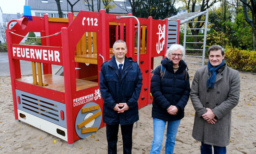 Drei Personen stehen auf einem Spielplatz, vor einem Spielgerät, das wie ein Feuerwehrauto aussieht