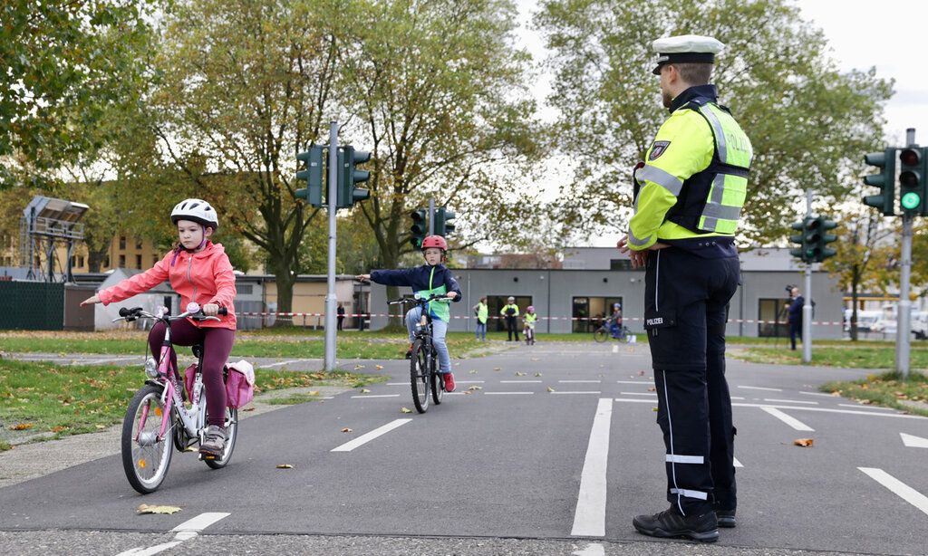 zwei Kinder fahren auf dem Verkehrsübugsplatz mit den Fahrrädern, Polizist guckt zu