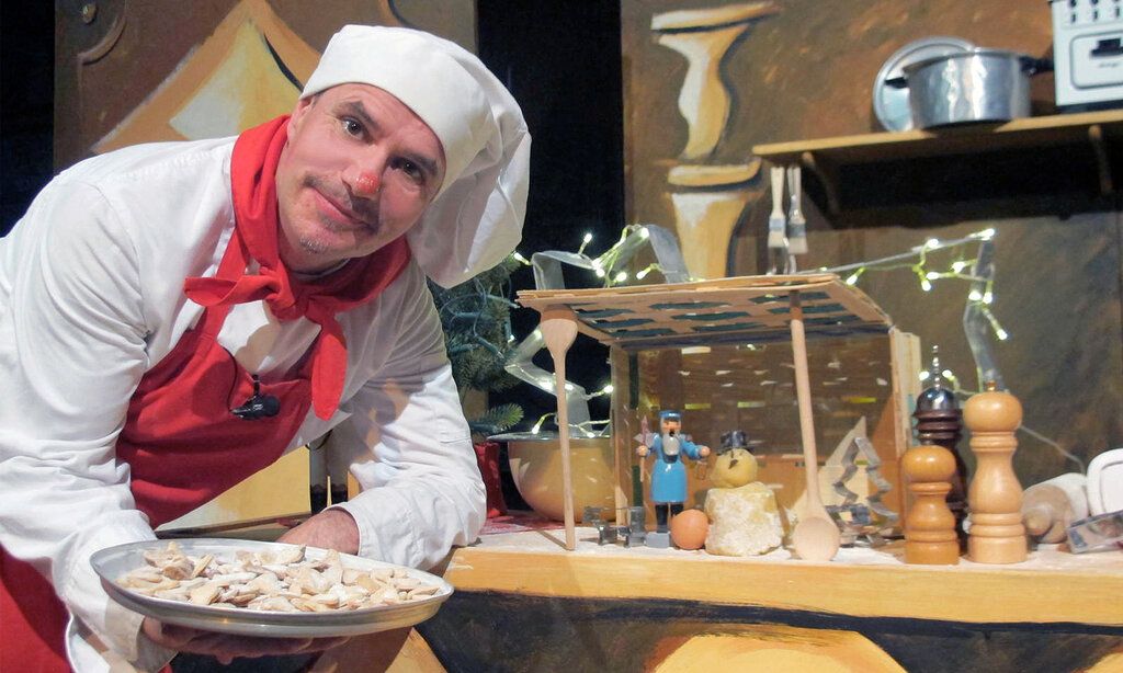 Mann mit Kochmütze und Schürze zeigt einen Teller mit Keksen, hinter ihm eine Küchen-Kulisse