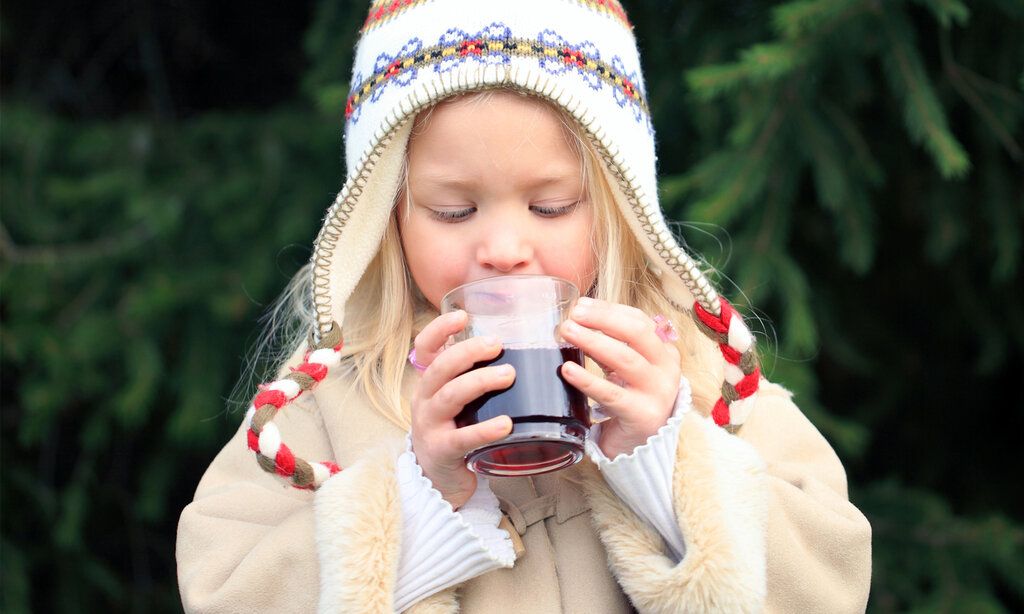 Winterlich gekleidetes Mädchen trinkt einen Kinderpunsch, im Hintergrund Tannen
