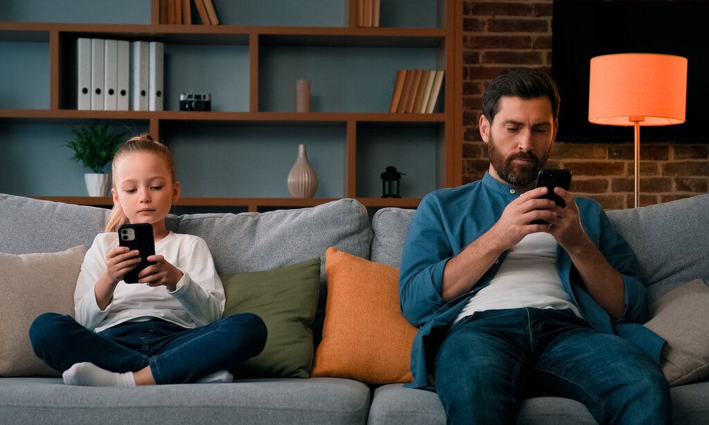 Tochter und Vater sitzen nebeneinander aufm Sofa, beide gucken auf ihr Smartphone