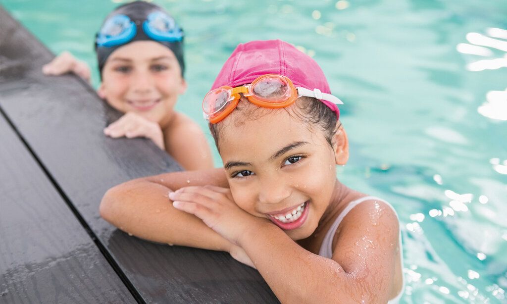 Ein Mädchen mit pinker Badekappe und Chlorbrille mit verschränkten Armen am Schwimmbeckenrand, im Hintergrund ein weiteres Kind