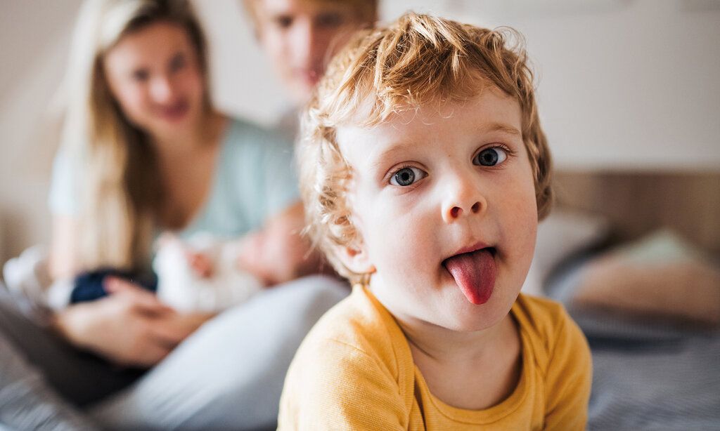 Kleiner Junge guckt in die Kamera und streckt die Zunge heraus, im Hintergund unscharf die Eltern mit einem Baby