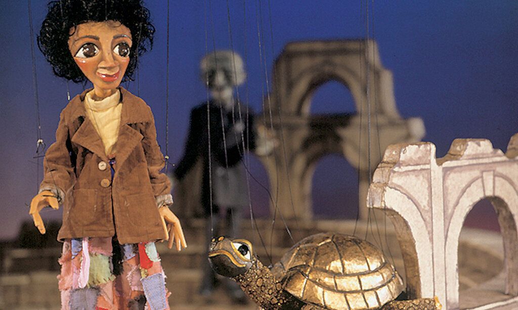 Marionette Momo steht mit einer goldenen Schildkröte auf der Bühne, im Hintergrund ein grauer Mann