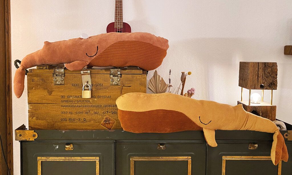 Zwei selbstgenähte Stillkissen in Form von Walen liegen auf großen Holzkisten, im Hintergrund eine Gitarre