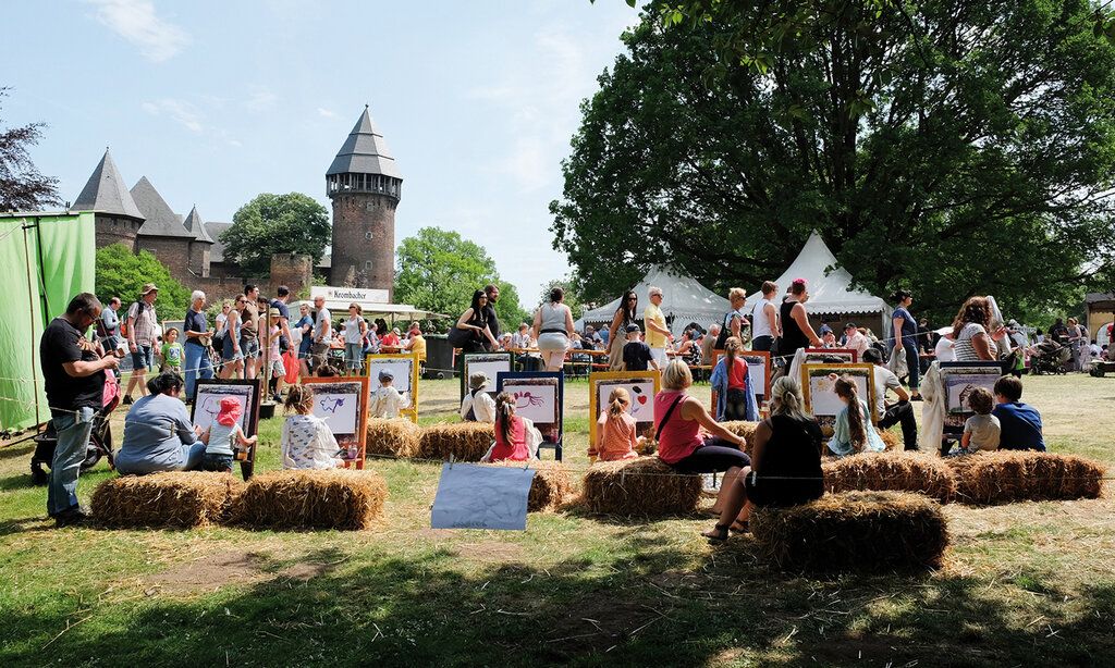 Leute sitzen auf Heuballen und malen Bilder, im Hintergund Marktstände und die Burg Linn
