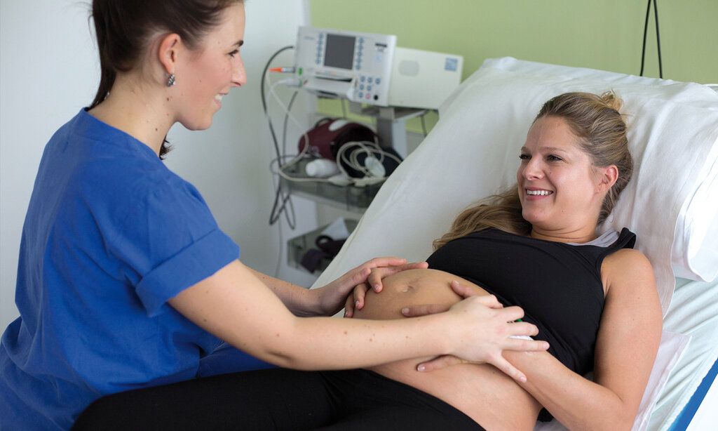 Schwangere Frau liegt im Krankenhausbett, eine Hebamme sitzt bei ihr, die Hände der beiden liegen auf dem Bauch der Schwangeren, beide lächeln