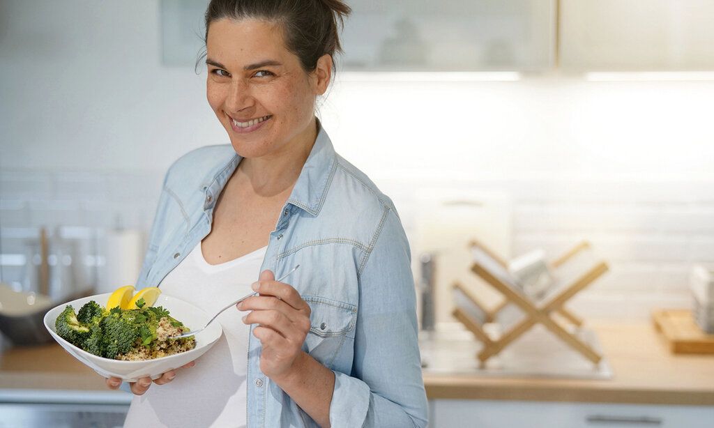 Schwangere Frau steht mit einem Teller, auf dem Brokkoli ist, in der Küche und lächelt in die Kamera