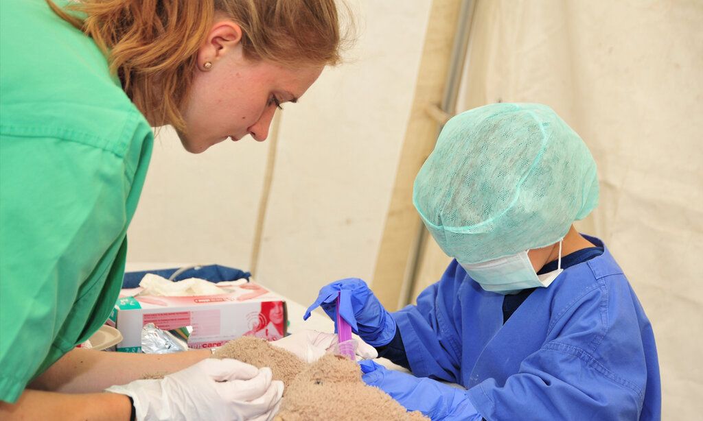 Kind in Arztkittel behandelt kranken Teddy, eine Frau assistiert