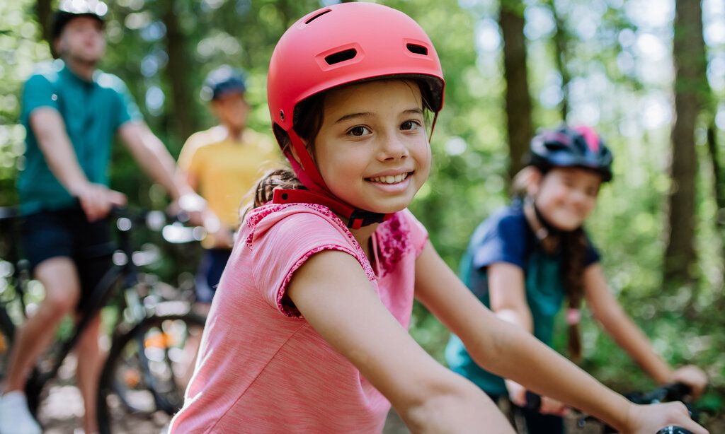 Mädchen mit rotem Fahrradhelm lächelt in die Kamera, im Hintergrund andere Radfahrer und Bäume, unscharf