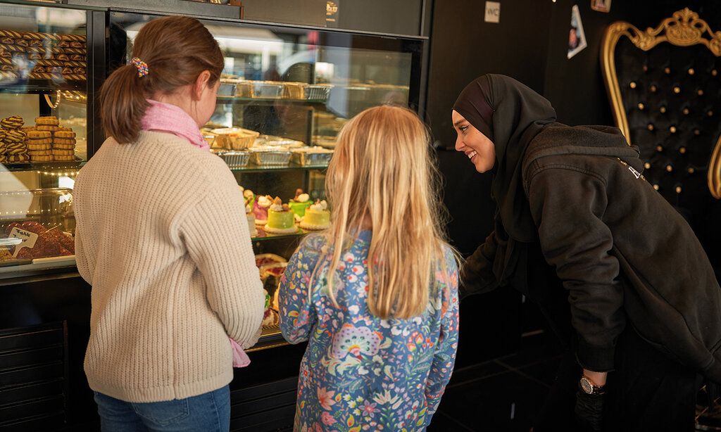 zwei Mädchen stehen im Cafe Hekimoglu und betrachten die Kuchen und Törtchen, eine Frau beträt sie.