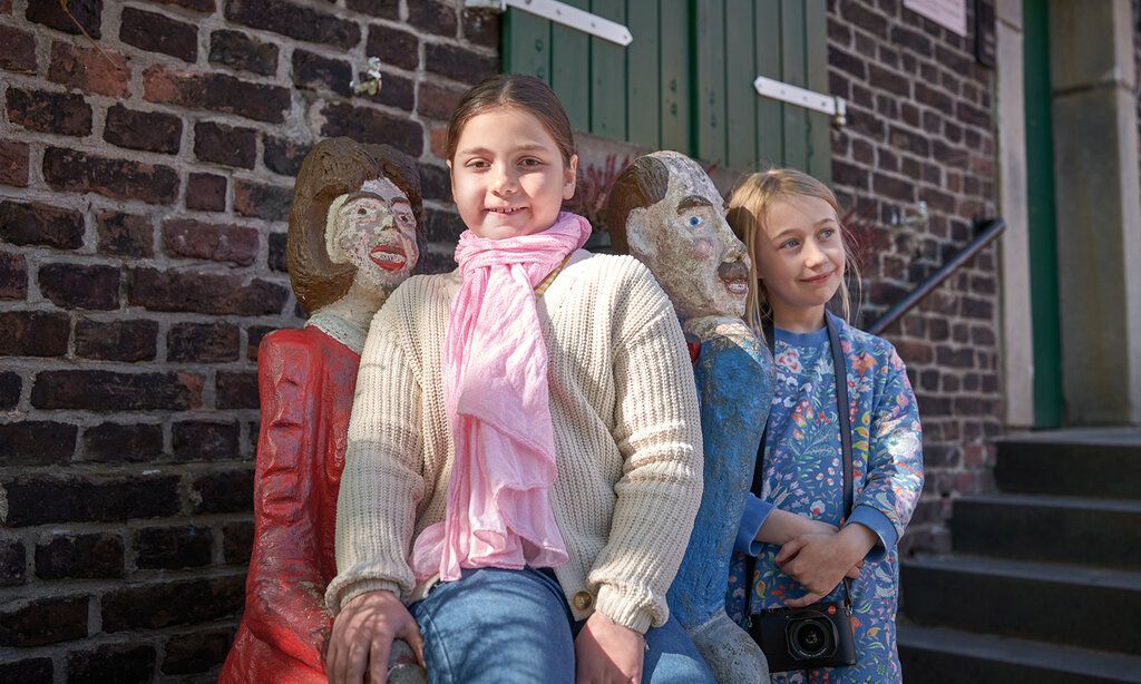Zwei Mädchen vor einer Hausmauer bei zwei Skulpturen, die eine Frau und einen Mann darstellen