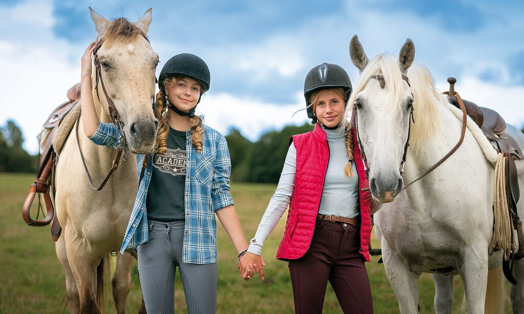 Zwei Mädchen mit Reiterhelmen stehen auf einer Wiese und halten sich an den Händen, neben ihnen jeweils ein Pferd
