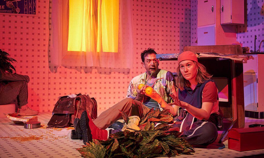 Eine Frau und ein Mann sitzen auf dem Boden der Bühne in der Kulisse einer Küche.