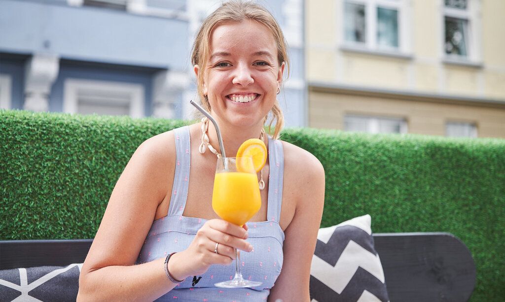 Eine junge Frau frühstückt im Café Liebe, lächelt in die Kamera undhält ein Glas Organgensfat vor sich