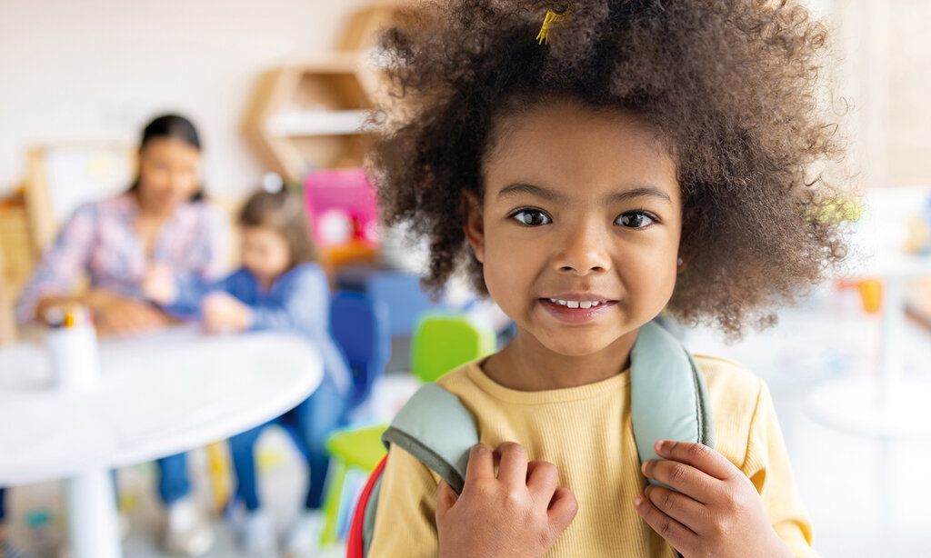 Porträt eines kleinen Schulmädchens, dahinter sitzt eine Lehrerin mit einer weiteren Schülerin an einem Tisch, Hintergrund unscharf